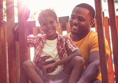 Father and son smiling together in a play area, with sunlight illuminating the scene.