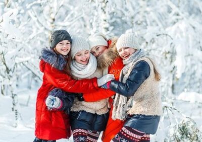 Four girls are standing together in a snowy forest, wearing warm winter clothing including hats and scarves. They are smiling, embracing each other, and appear joyful in the wintry scene filled with snow-covered trees.