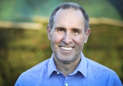 A smiling middle-aged man with short, graying hair is wearing a light blue button-up shirt. He is outdoors, with soft, muted greenery in the background that suggests a calm and pleasant setting.