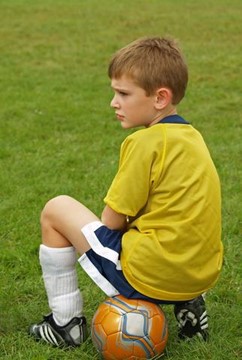 A boy in a yellow soccer jersey sits on a ball, looking thoughtful on a grassy field.