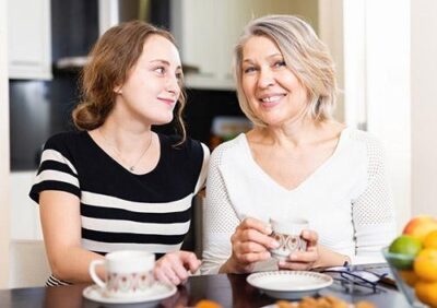 A young woman and an older woman sit together, smiling, with tea cups on a table filled with fruit.